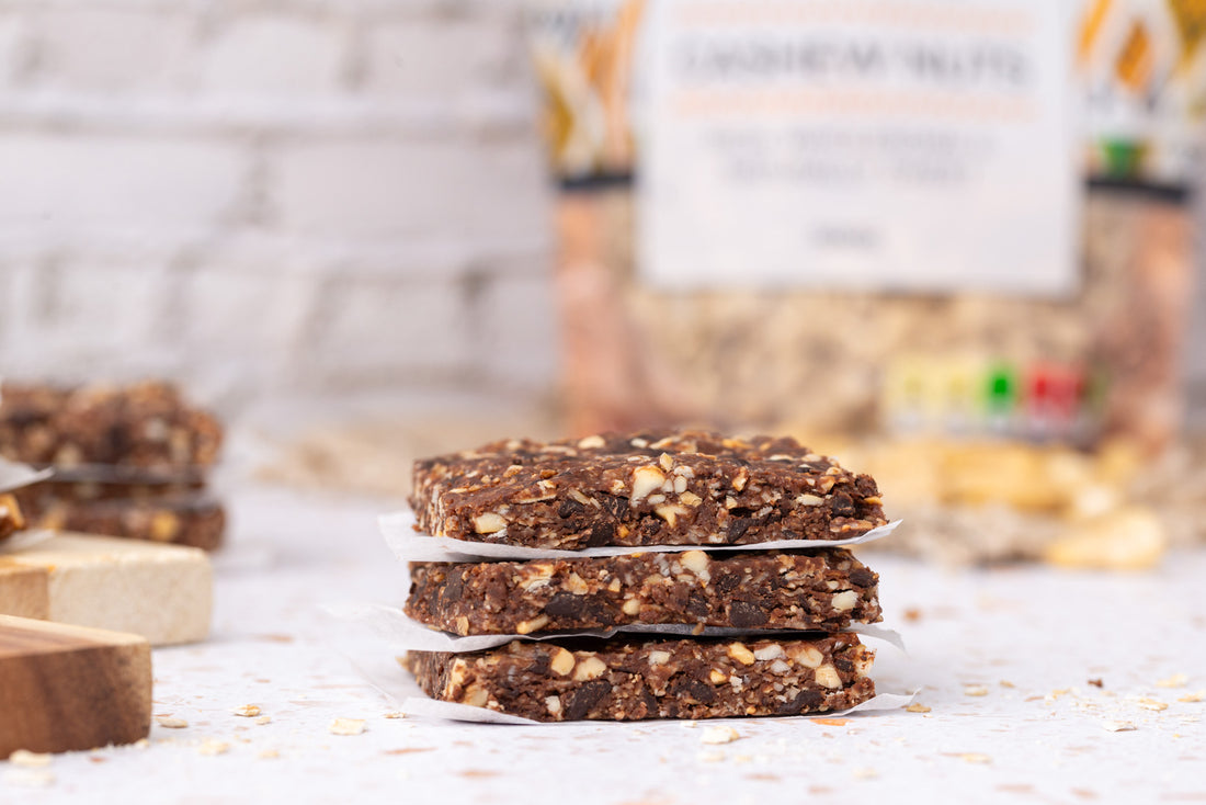 Three homemade Chocolate Cashew Energy Bars stacked atop one another, with a bag of Essina Wholefoods raw cashew nuts visible in the background, showcasing the key ingredient in these nutritious and delicious homemade energy bars